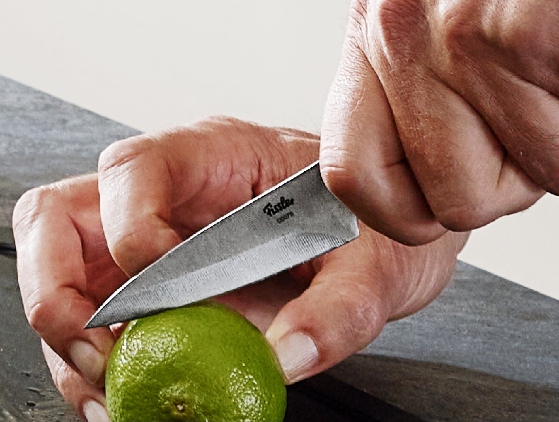Eine Nahaufnahme von Händen, die eine grüne Limette auf einer dunklen Oberfläche mit einem Fissler-Messer schneiden, das deutsche Präzision und zeitloses Design demonstriert.