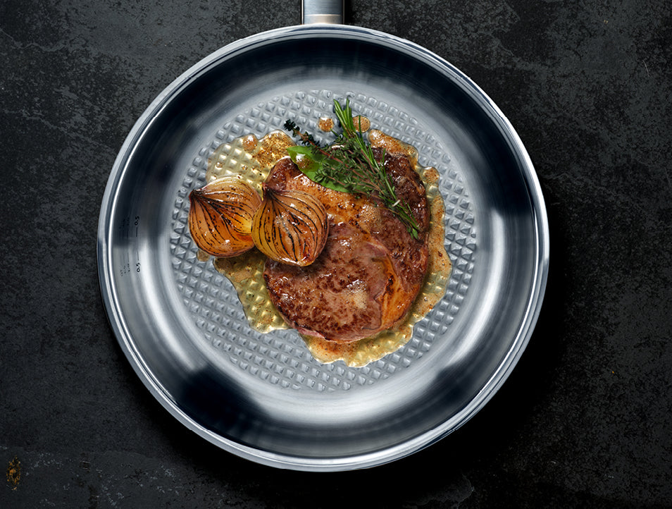 Ein perfekt gebratenes Steak mit Zwiebeln, Rosmarin und Sauce in einer Fissler-Edelstahlpfanne auf einer dunklen Arbeitsplatte.