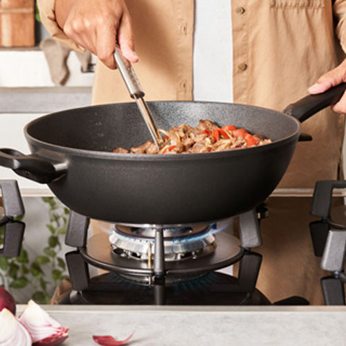 A person cooks with a Fissler Adamant® wok, known for German precision, on a gas stove as fresh ingredients sizzle.