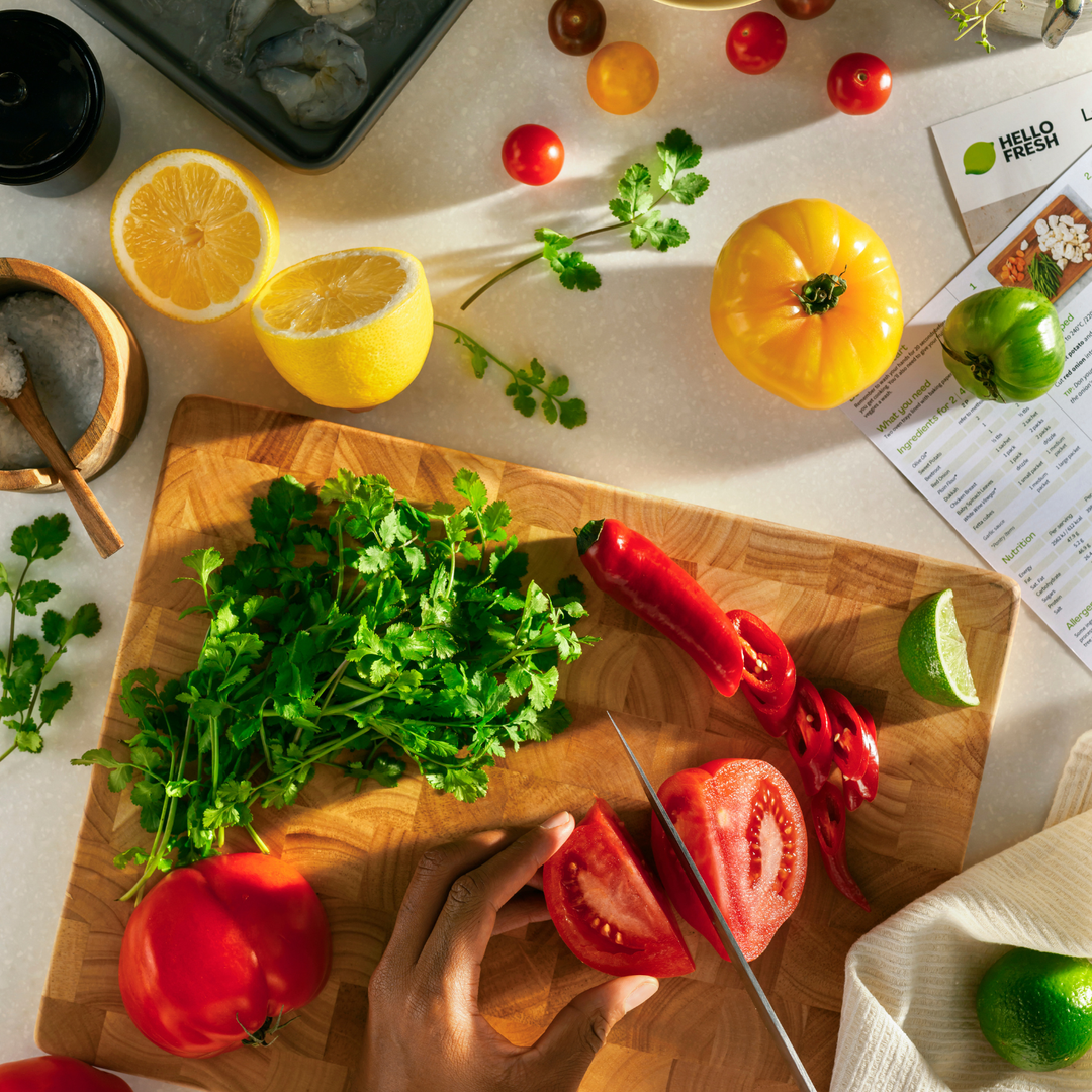 Eine Hand schneidet gekonnt eine reife rote Tomate auf einem Holzschneidebrett, umgeben von frischem Koriander, roten Chilischoten, Limetten, gelben Tomaten, Zitronen und Kirschtomaten. Die Szene spiegelt eine sorgfältige Zubereitung wider, bei der präzise Küchenutensilien zum Einsatz kommen, die an das in Deutschland entwickelte Kochgeschirr von Fissler und eine zeitlose kulinarische Tradition erinnern.