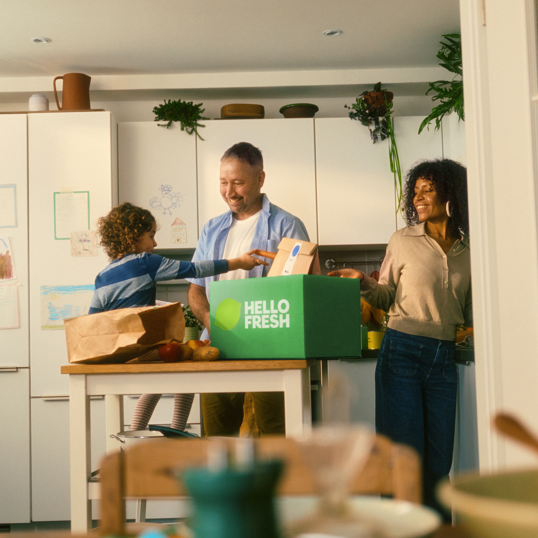 Eine dreiköpfige Familie versammelt sich in einer hellen Küche und packt gemeinsam lächelnd frische Zutaten aus, während auf der Arbeitsplatte eine Auswahl an hochwertigem Fissler-Edelstahl-Kochgeschirr ausgestellt ist. Auf den weißen Schränken im Hintergrund sind Zeichnungen zu sehen, die dem zeitlosen Raum, der durch präzise gefertigte Töpfe und Pfannen „Made in Germany“ geprägt ist, eine warme Atmosphäre verleihen.