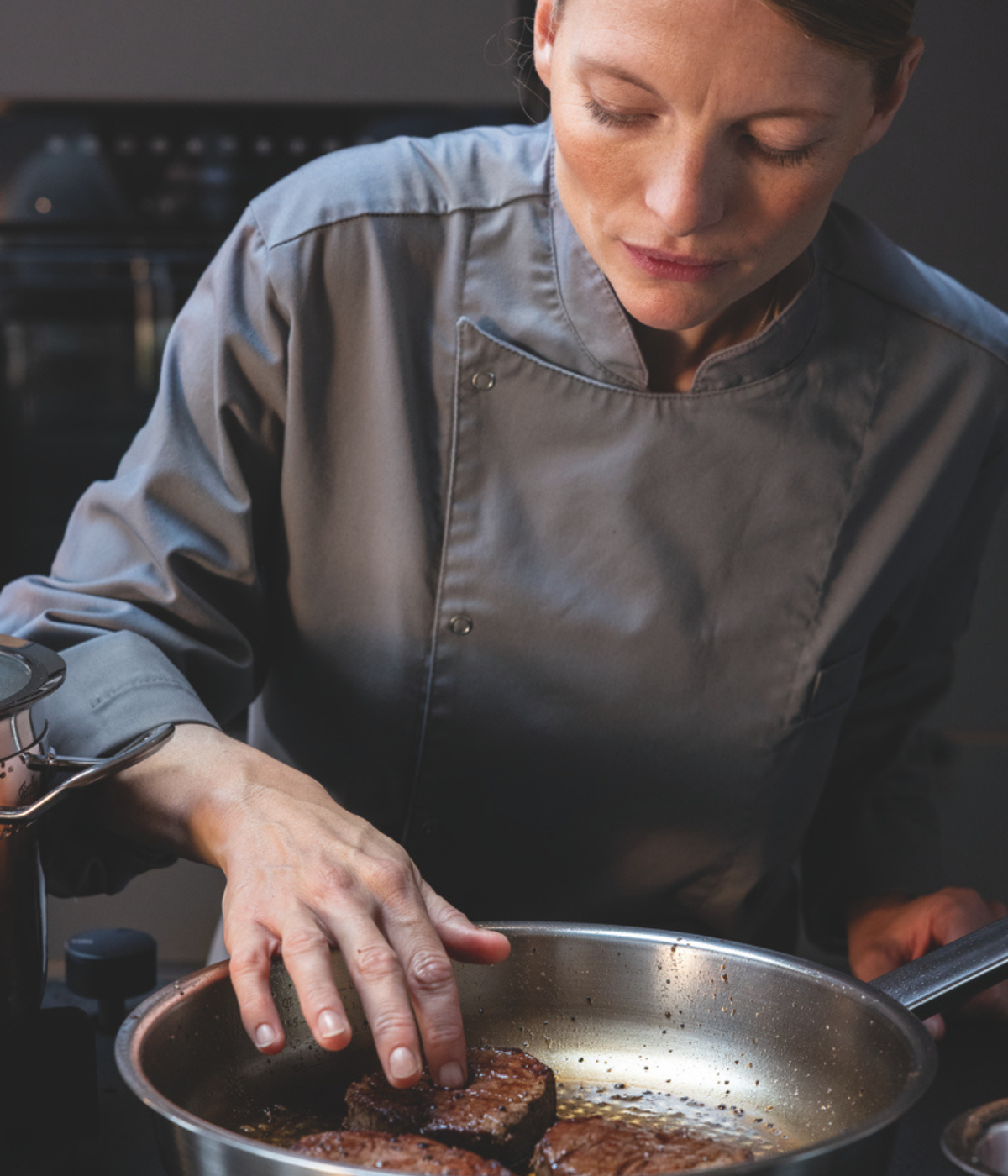 Ein Koch in grauer Uniform prüft sorgfältig den Gargrad eines Steaks, das in einer hochwertigen Fissler-Edelstahlpfanne brutzelt, und unterstreicht damit die Präzision und Qualität der in Deutschland entwickelten Fissler-Kochgeschirre.