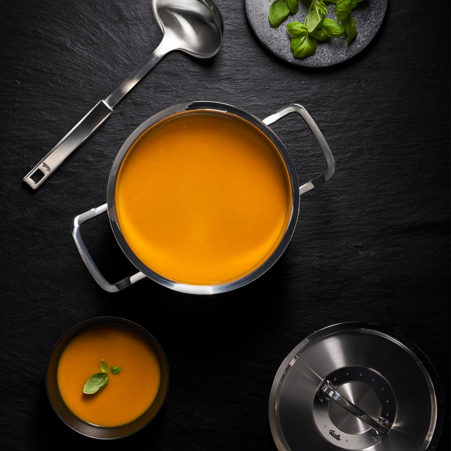 Fissler stainless steel pot with creamy soup on a dark surface, lid and ladle next to it; bowl with soup, garnished with basil.
