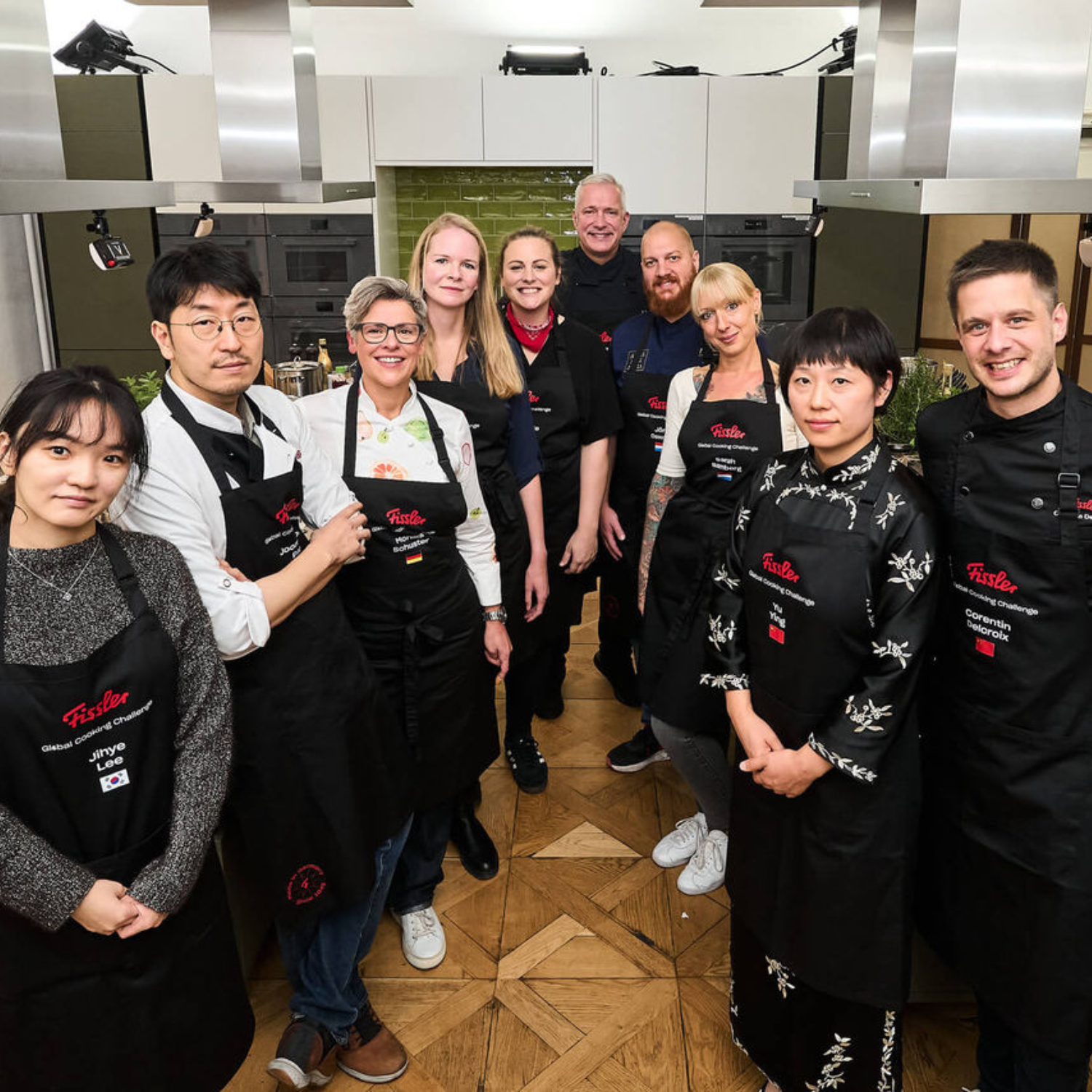 Zehn Personen in schwarzen Schürzen lächeln in einer eleganten Küche mit Fissler-Edelstahlkochgeschirr zusammen und strahlen Teamwork und Innovation aus.