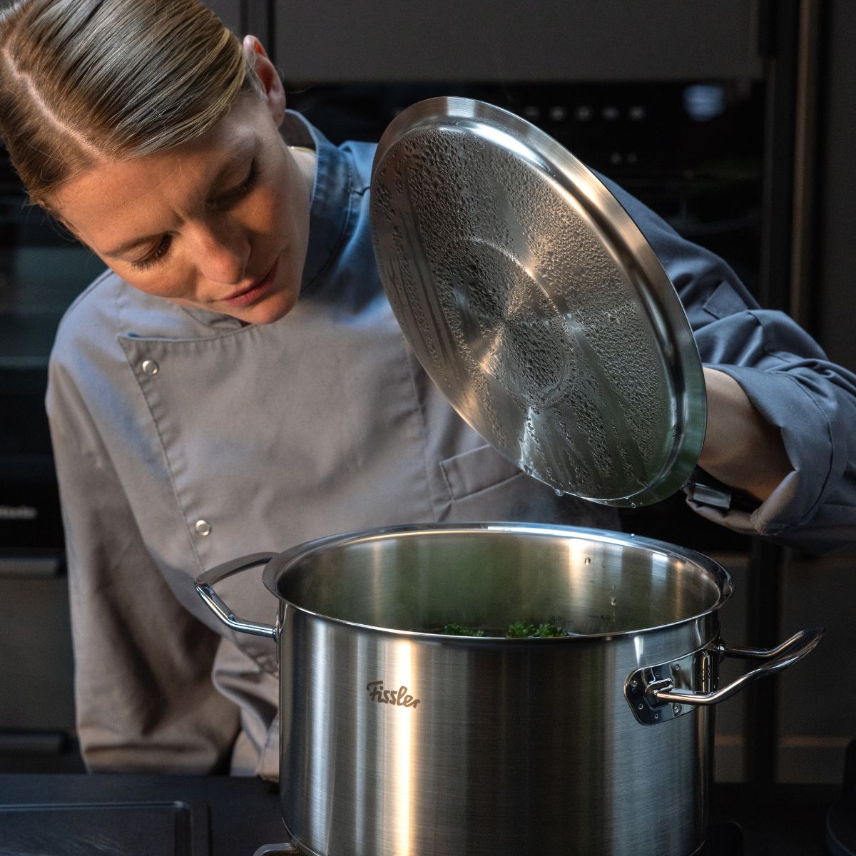 A chef in a gray coat lifts the lid of a Fissler stainless steel pot on the stove, cooking with German precision and design.