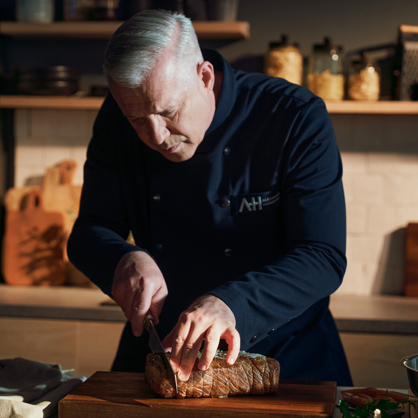 Ein Koch mit grauen Haaren schneidet einen Braten auf einem Holzbrett mit dem Präzisionsmesser von Fissler, im Hintergrund stehen Kochgeschirr und Töpfe.