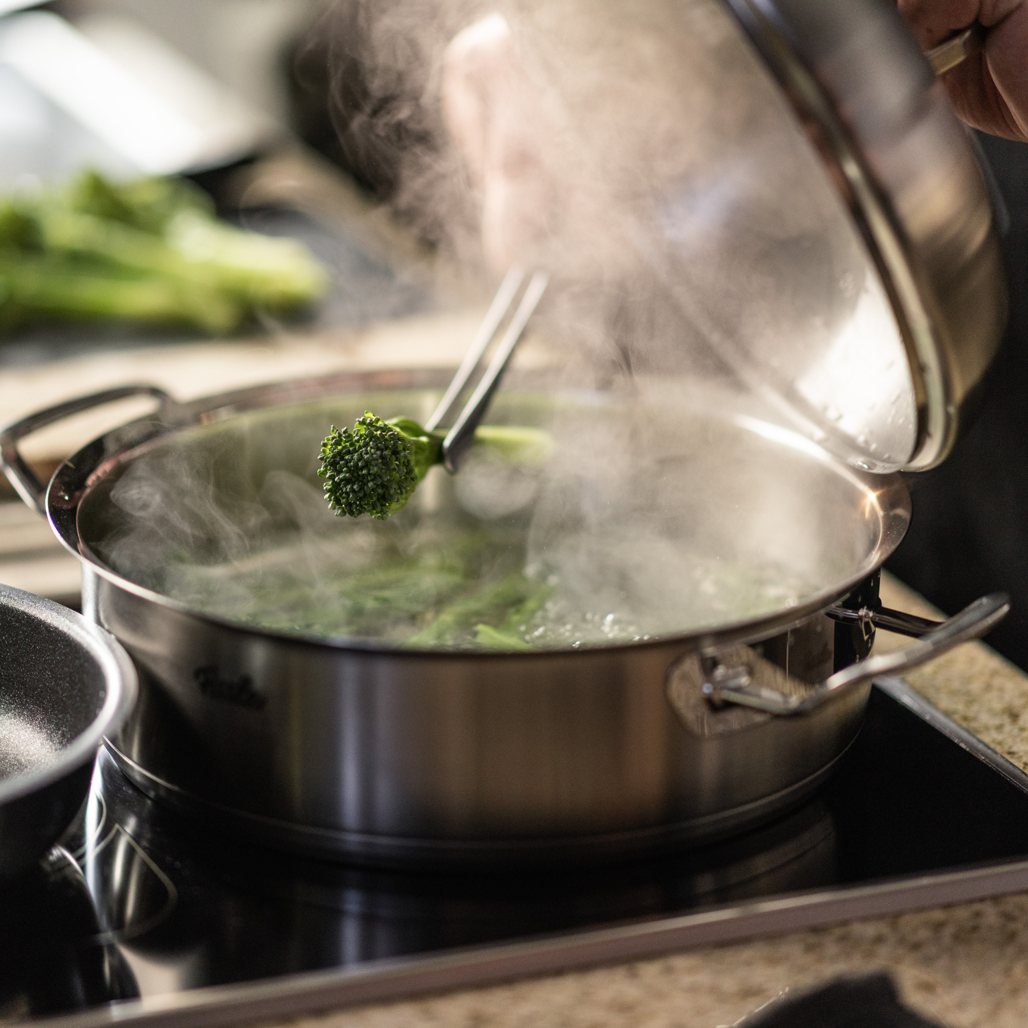 Ein Koch öffnet einen Fissler-Edelstahltopf, der auf dem Herd dampft, und hält mit einer Zange Brokkoli fest. Frisches Gemüse unterstreicht die Qualität der Küche.