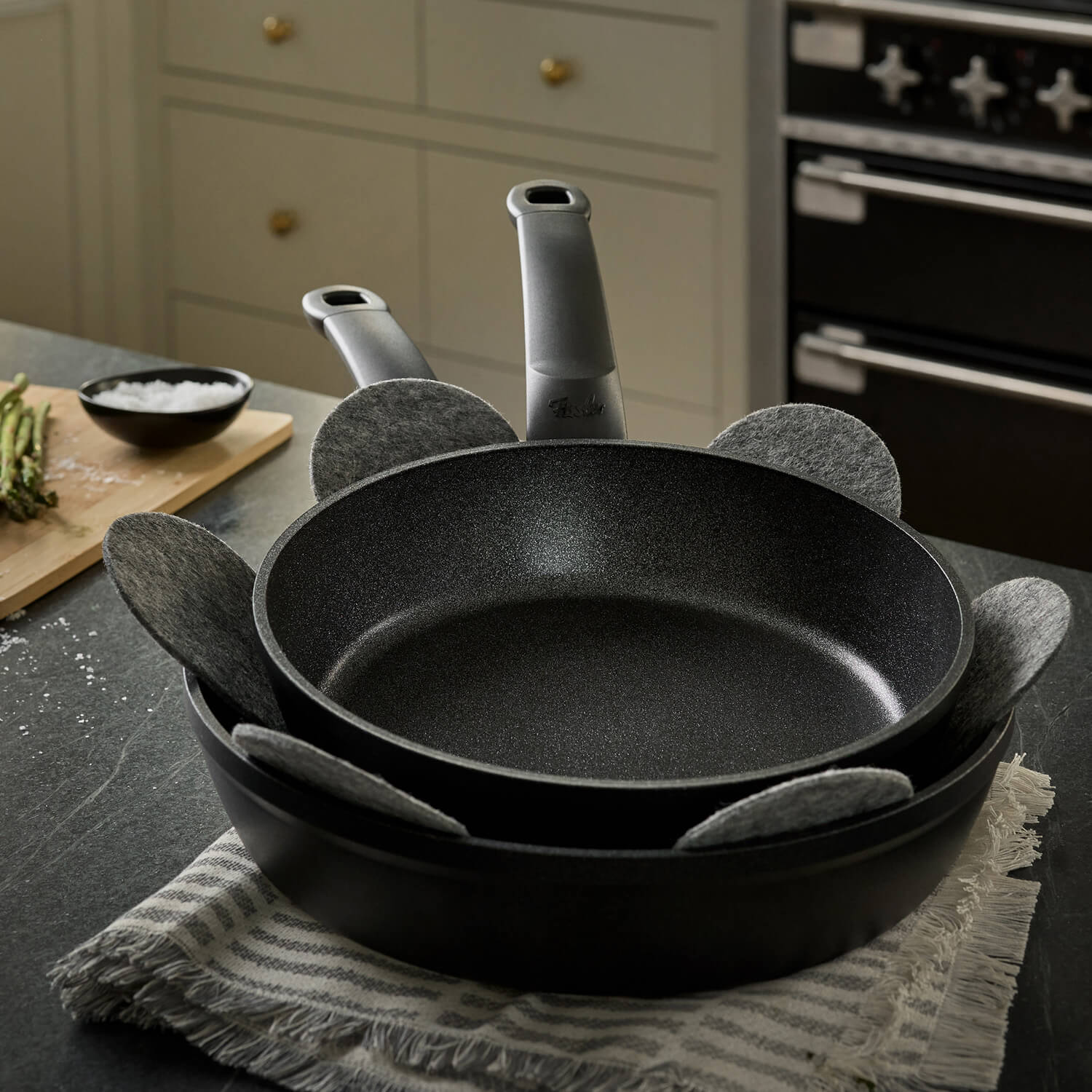 Zwei Fissler Antihaft-Pfannen mit grauem Pfannenschutz, gestapelt auf einem gestreiften Tuch, sind Ausdruck deutscher Innovation und zeitlosen Designs.