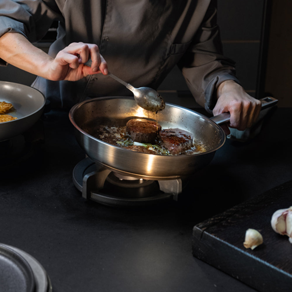 A chef expertly cooks steaks in a premium Fissler stainless steel pan on a stovetop, carefully spooning sauce over the meat. Nearby, fresh garlic and a stylish dark cutting board complement the professional kitchen scene, showcasing Fissler’s commitment to precision engineering and timeless design.
