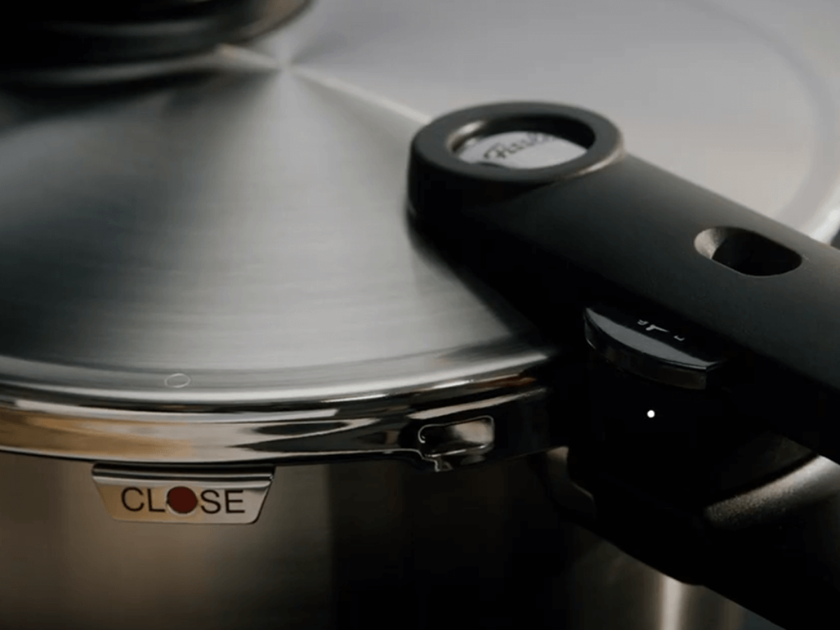 Close-up of Fissler pressure cooker lid with polished stainless steel, ergonomic handle, signature knob, and CLOSE label in red and black.