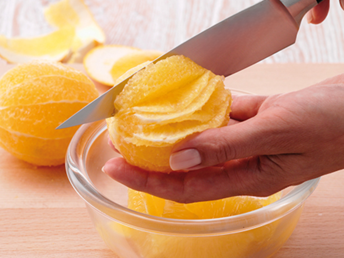 Eine Hand schneidet mit Präzision eine geschälte Orange über einer gläsernen Fissler-Schale; die Orangenschalen liegen auf einer Holzfläche im Hintergrund.