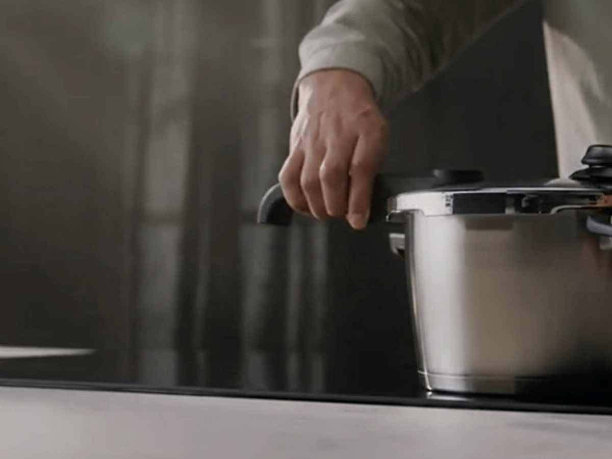 Eine Hand hält den ergonomischen Griff eines Fissler-Edelstahl-Schnellkochtopfs, der für deutsche Präzisionstechnik und zeitloses Design bekannt ist, während dieser auf einer eleganten, dunklen Küchenarbeitsplatte steht.