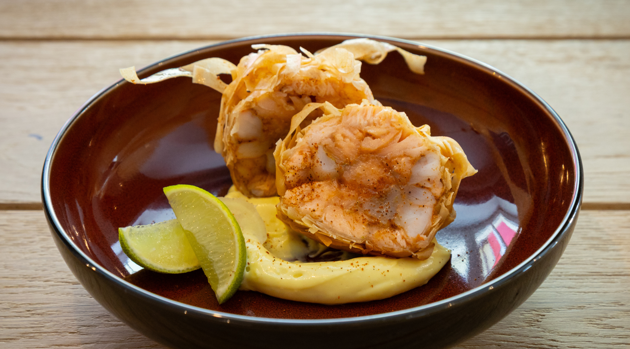 Two pieces of crispy seafood wrapped in golden pastry are elegantly arranged on a dark plate with creamy sauce, accompanied by two lime wedges. Presented on a light wooden surface, this dish highlights the precision and quality you can achieve with Fissler’s premium stainless steel cookware—engineered for durability, timeless design, and innovation made in Germany.