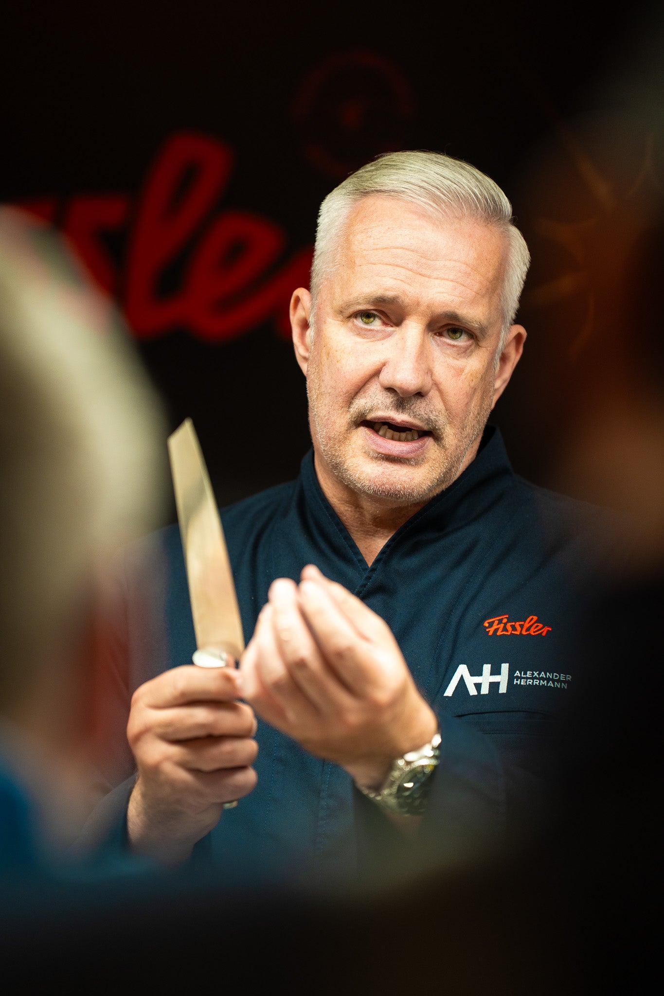 Ein Koch in einer dunkelblauen Jacke zeigt ein Fissler-Messer und gestikuliert, während er spricht. Unscharfer Hintergrund und rotes Fissler-Branding im Hintergrund.