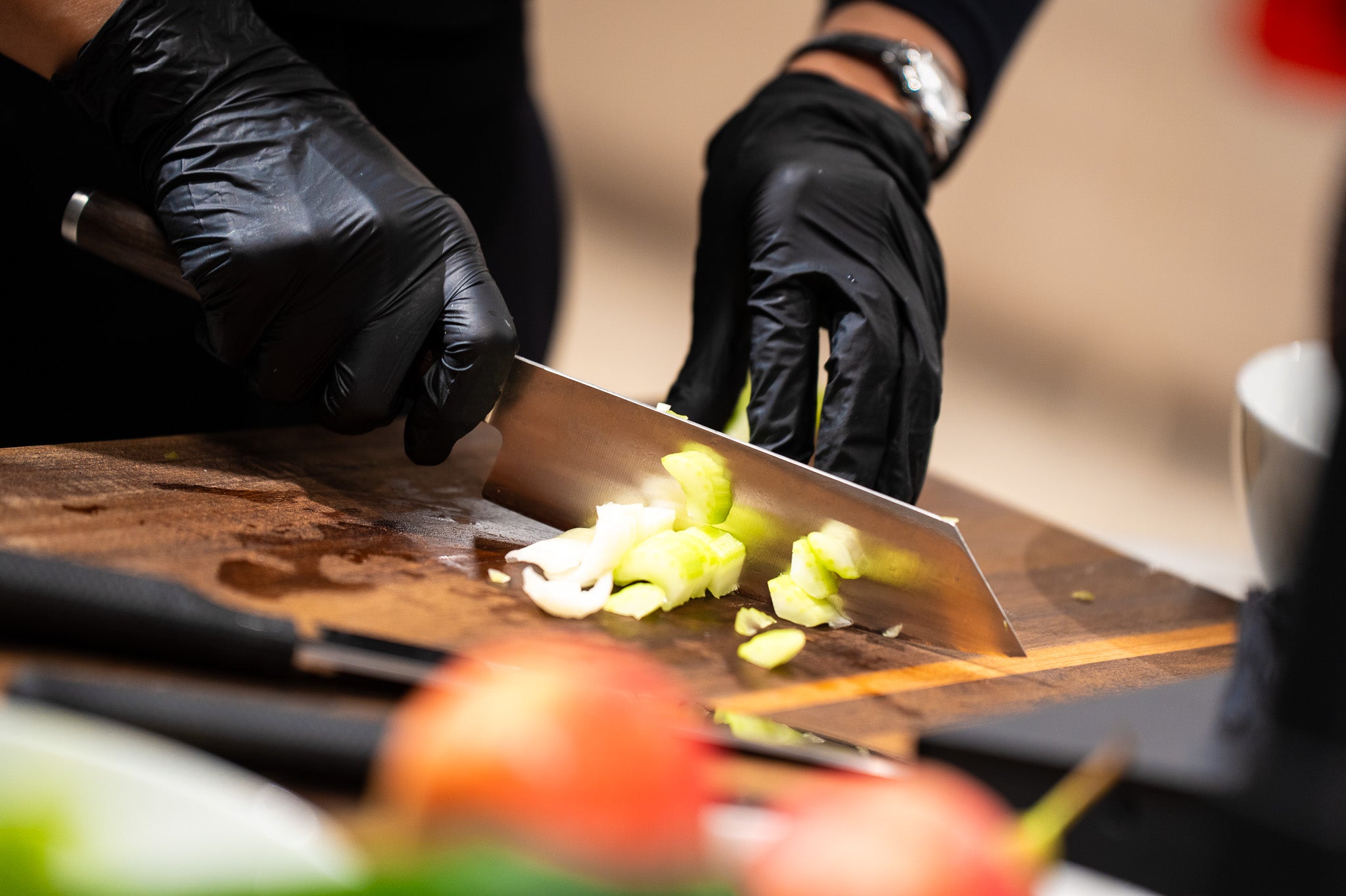 Eine Person mit schwarzen Handschuhen schneidet Sellerie auf einem Holzbrett mit einem Fissler-Kochmesser. Im Vordergrund steht hochwertiges Kochgeschirr.