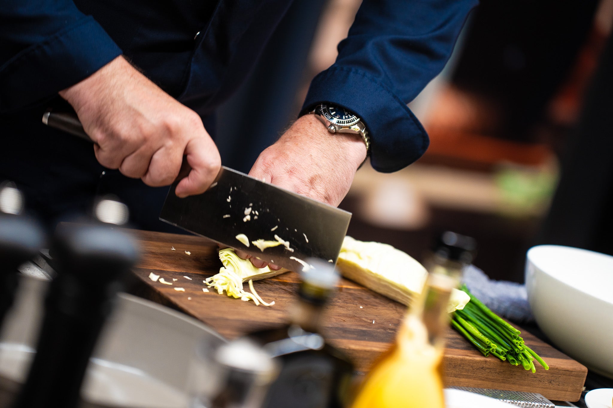 Eine Person in einem dunkelblauen Hemd schneidet Gemüse mit einem Fissler-Messer auf einem Holzbrett, daneben Fissler-Kochgeschirr und grüne Zwiebeln.