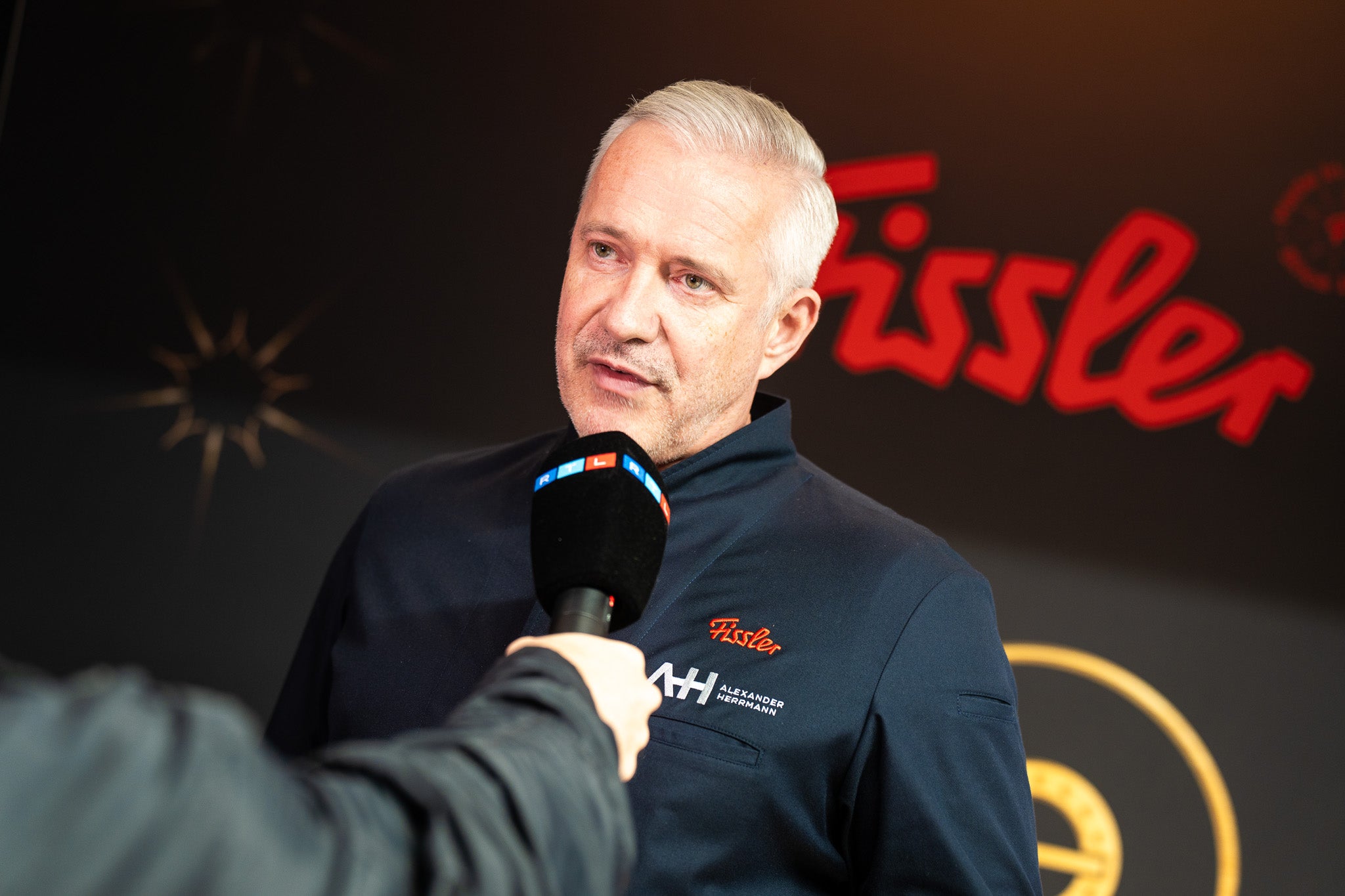 Ein Mann in einer Fissler-Jacke wird interviewt, hinter ihm das ikonische rote Logo, das seit 1845 für innovatives deutsches Kochgeschirr steht.