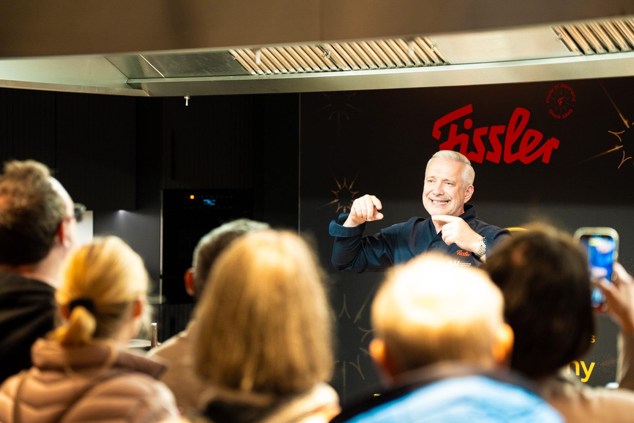 Ein lächelnder Mann steht vor einem Fissler-Schild und präsentiert dem Publikum, während die Gäste das hochwertige Kochgeschirr im Fokus haben.
