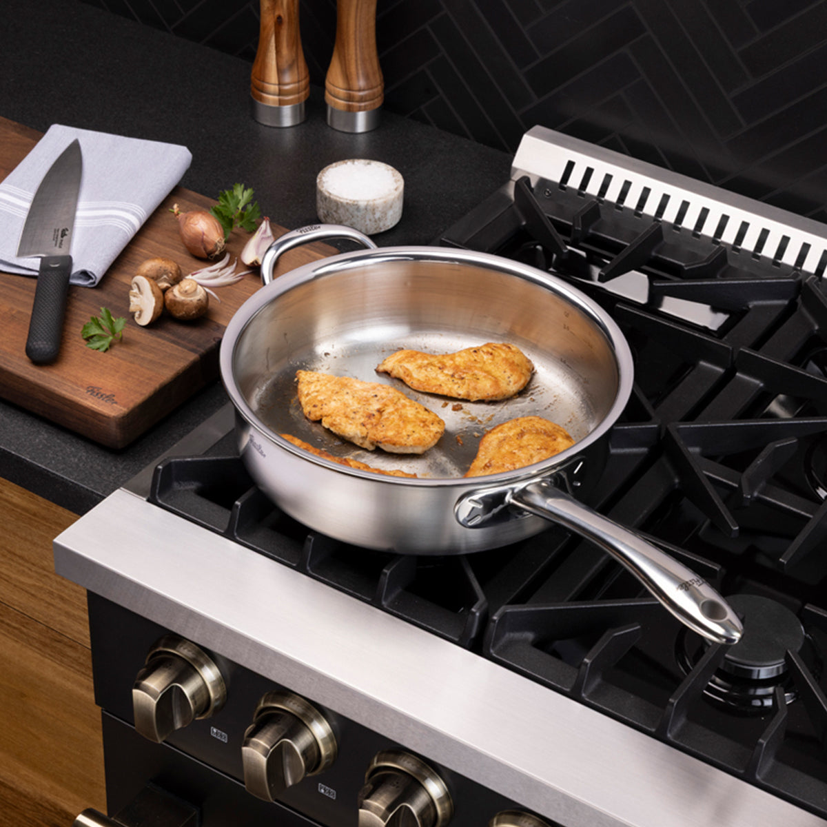 Three pieces of chicken are being expertly cooked in the Fissler M5 Pro-Ply® 3-piece set with metal lid on a gas stove. Nearby, a cutting board holds a knife, fresh mushrooms, garlic, aromatic herbs, and a towel. The high-quality induction-compatible Fissler cookware—precision-engineered in Germany since 1845 and renowned for durability and timeless design—stands out against the sleek modern black kitchen backsplash.