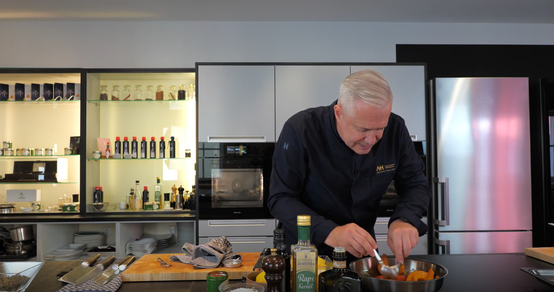 A chef with gray hair uses Fissler stainless steel cookware to mix ingredients in a modern kitchen, with bottles and shelves in the background.