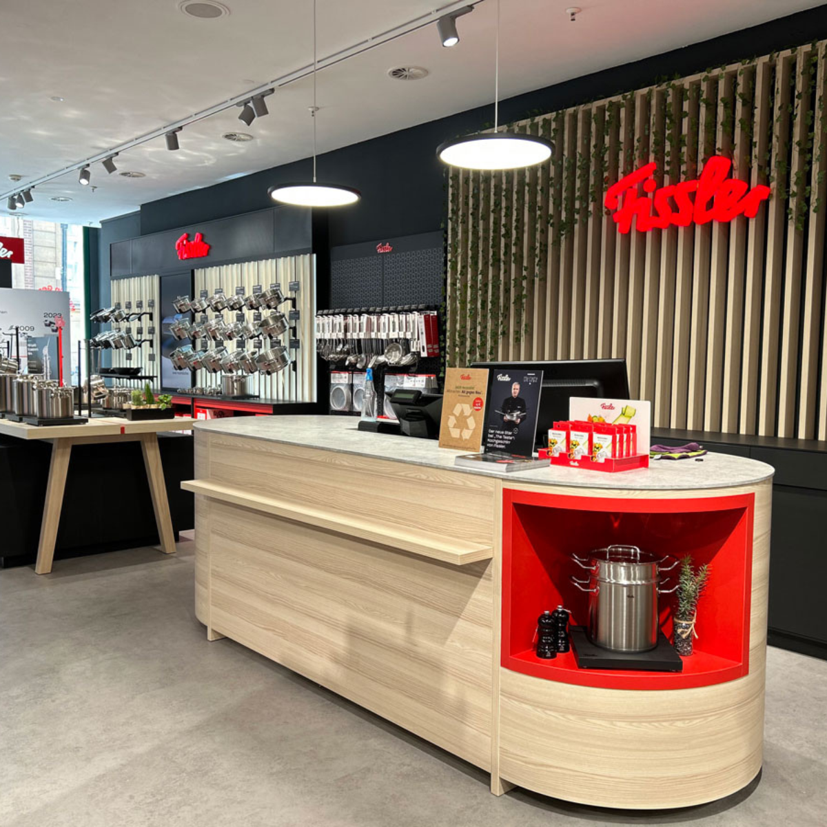 Fissler Ladeneinrichtung mit hellem Holztresen, roten Regalen mit Kochgeschirr aus deutscher Produktion, Töpfen und Pfannen aus Edelstahl.