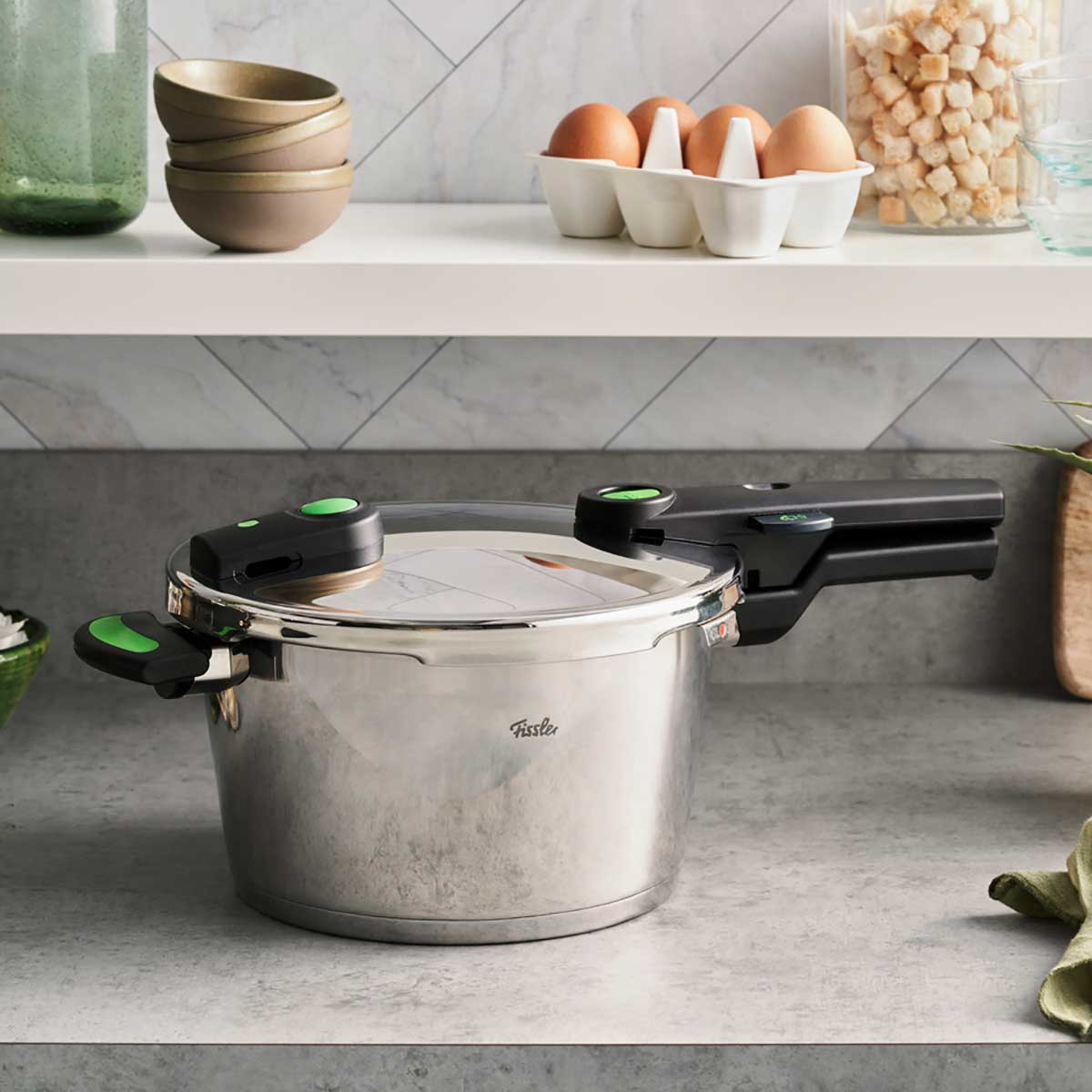 Fissler Vitaquick® Green pressure cooker with distinctive handles on a gray plate, surrounded by kitchen accessories.