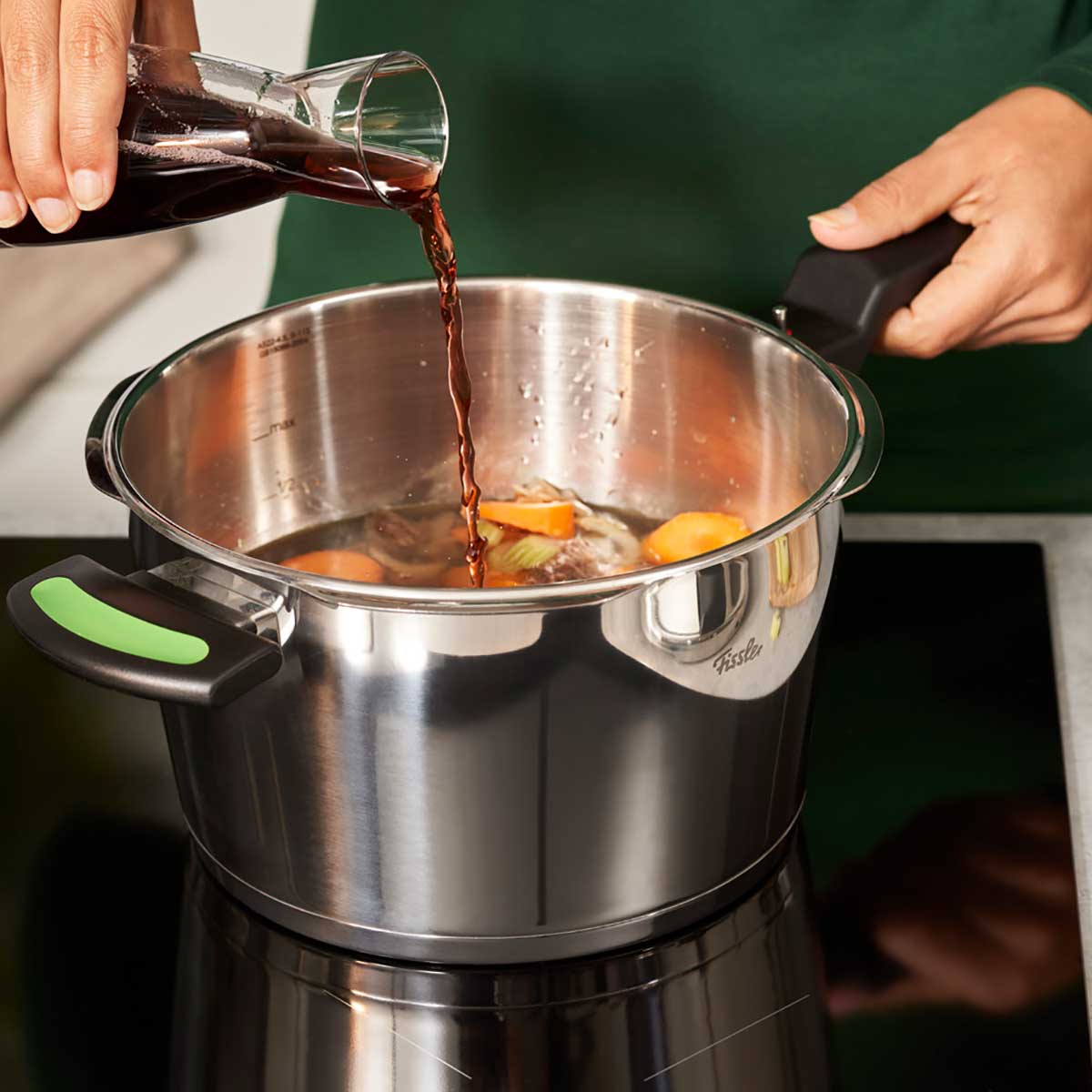 A hand pours a dark liquid into vegetables simmering in the Fissler Vitaquick® Green pressure cooker for healthy cooking.