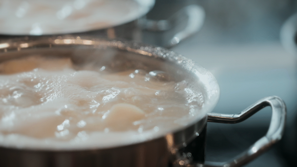 Nahaufnahme aus der Vogelperspektive von zwei hochwertigen Fissler-Edelstahltöpfen auf einem Herd, die jeweils mit sprudelndem Wasser und perfekt geschnittenen Kartoffeln gefüllt sind. Der Dampf steigt elegant über dem präzise gefertigten Kochgeschirr auf und erzeugt einen Weichzeichner-Effekt, der die Langlebigkeit und das zeitlose deutsche Design unterstreicht.