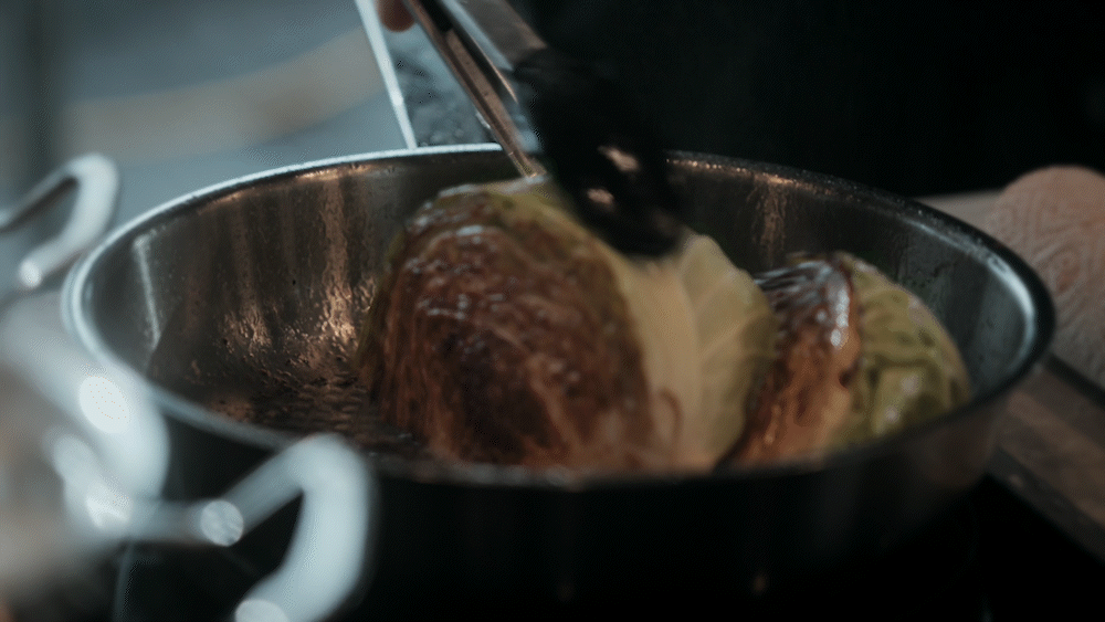 Eine Person brät einen halbierten Kohlkopf in einer Fissler-Edelstahlpfanne mit einer Zange an, wobei die Schnittflächen gebräunt werden. Die hochwertige deutsche Technik der Pfanne sorgt für eine gleichmäßige Wärmeverteilung und perfekte Ergebnisse.