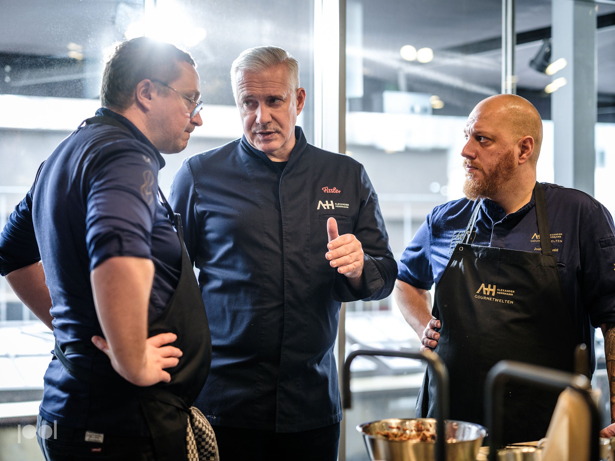 Three chefs in dark uniforms stand indoors, engaged in an intense discussion, reflecting precision and dedication. One chef gestures purposefully as the others listen attentively. Stainless steel Fissler cookware, including pots, pans, and premium kitchen accessories designed for exceptional performance and durability, are visible alongside fresh ingredients in the foreground.
