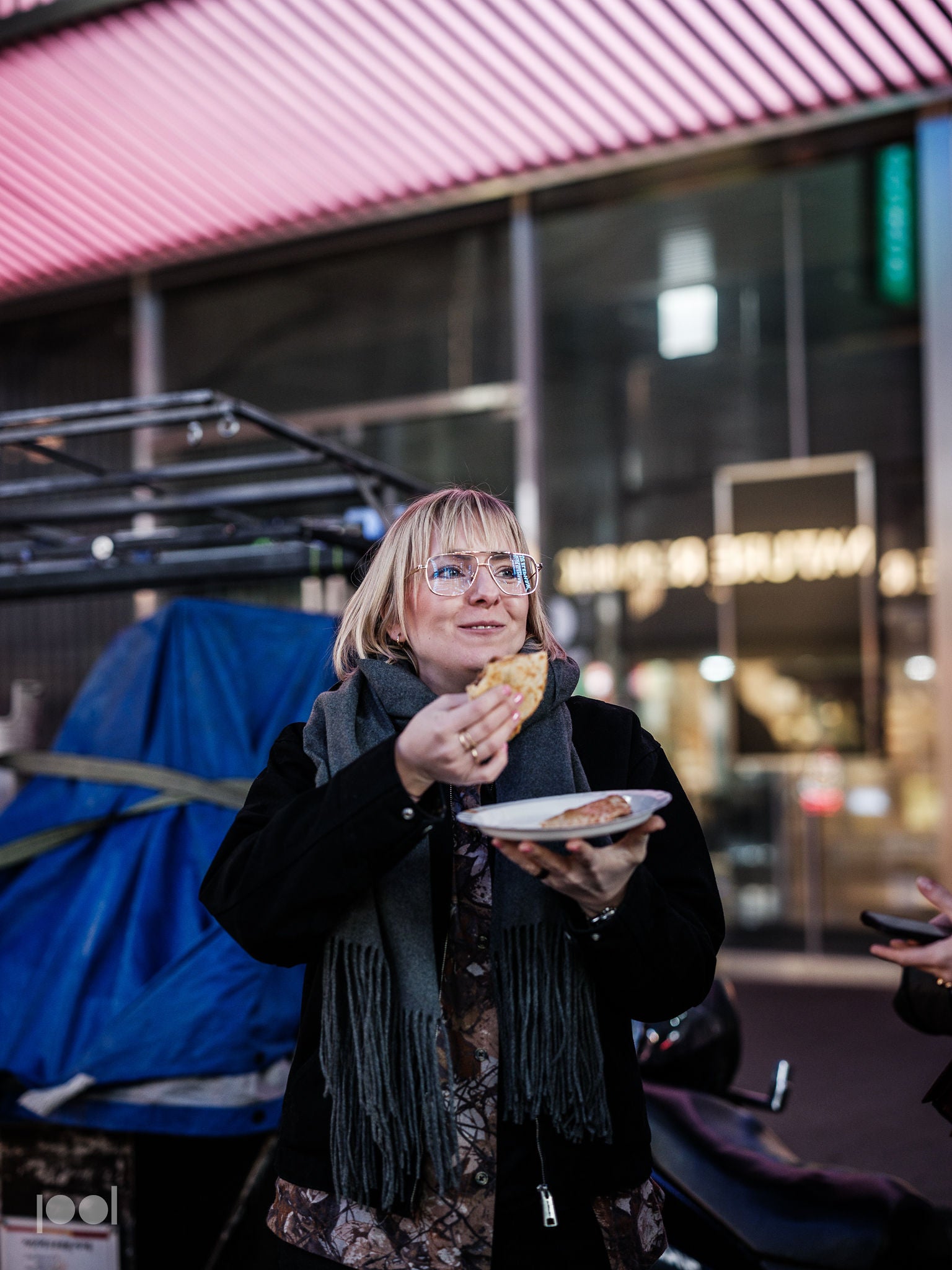 Eine Frau mit Brille und Schal lächelt, während sie im Freien in einer städtischen Umgebung, die von Neonlichtern und verschwommenen Ladenschildern beleuchtet wird, eine Mahlzeit genießt, die auf einem Teller serviert wird. Die Szene spiegelt die Freude am Essen mit präzise gefertigtem Kochgeschirr wider, inspiriert von der Fissler-Tradition von Qualität, Langlebigkeit und zeitlosem deutschem Design.